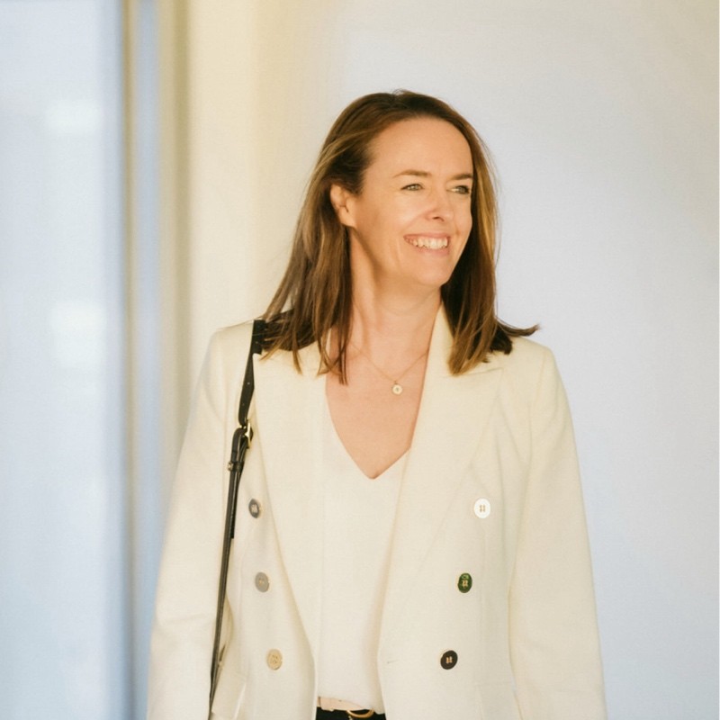 Bridget O'Neill, smiling and looking off to one side, wearing a cream blazer against a pale wall.
